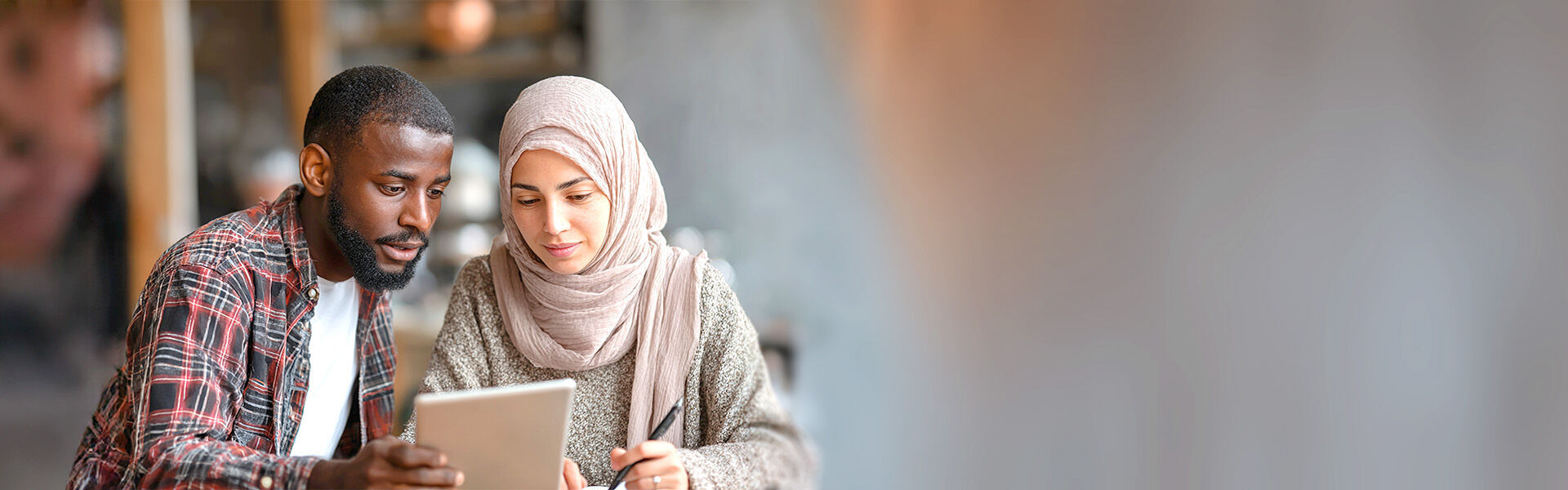 Junge Frau mit Kopftuch und ein dunkelhäutiger junger Mann sitzen in einem Großraumbüro und schauen gemeinsam auf ein Tablet.